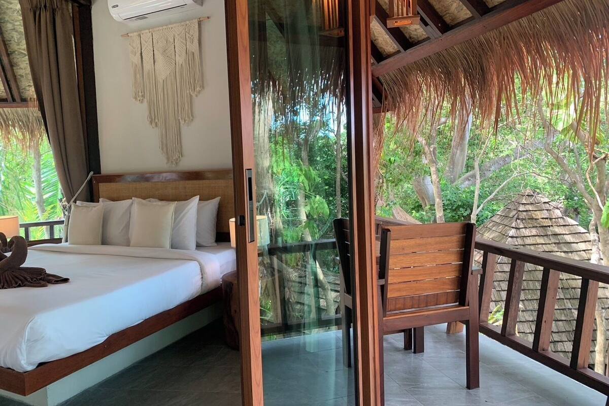 A bedroom with a bed, a bedside table, and a chair. There is a window with a view of trees and a thatched roof structure outside.