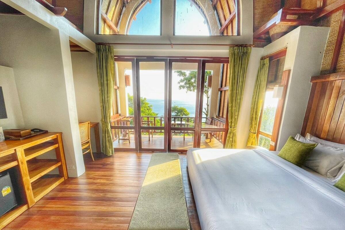 A bedroom with a large bed, a wooden desk, and a balcony with a view of the ocean.