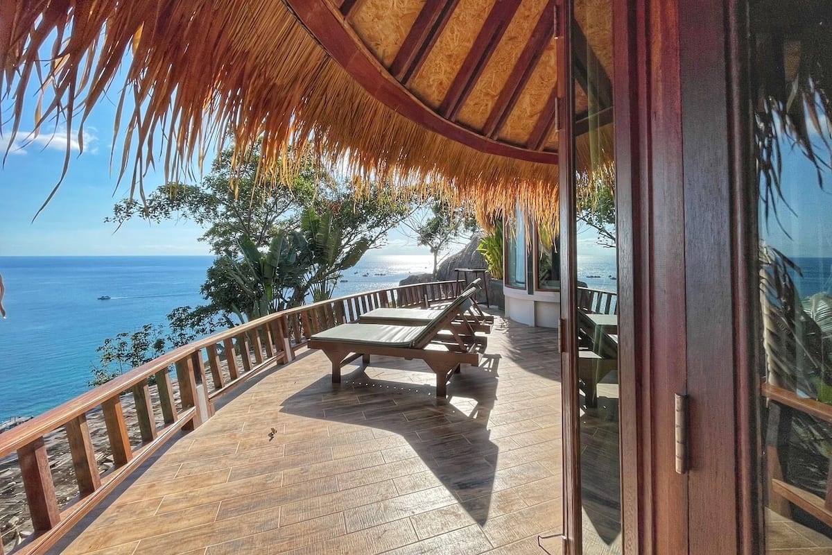 A wooden deck with a thatched roof, overlooking a beach and the ocean.