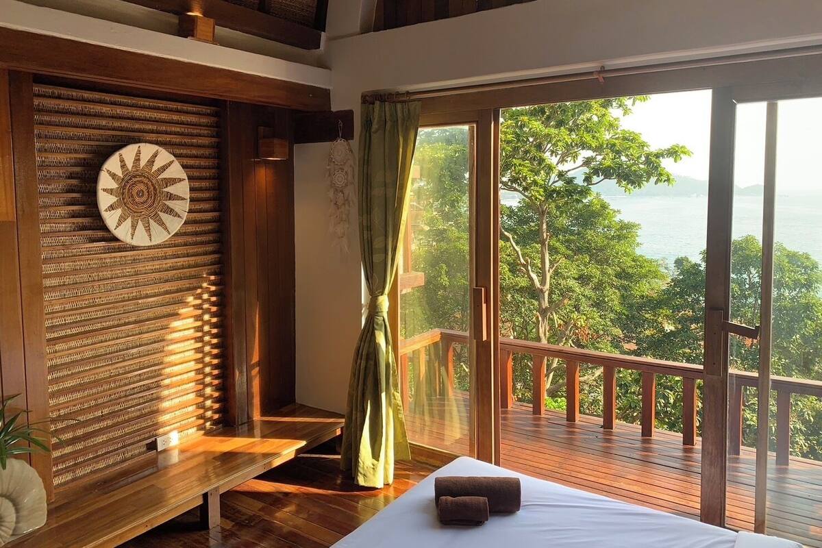 A room with a bed, a bench, a wall with a circular design, and a view of a balcony with a railing.