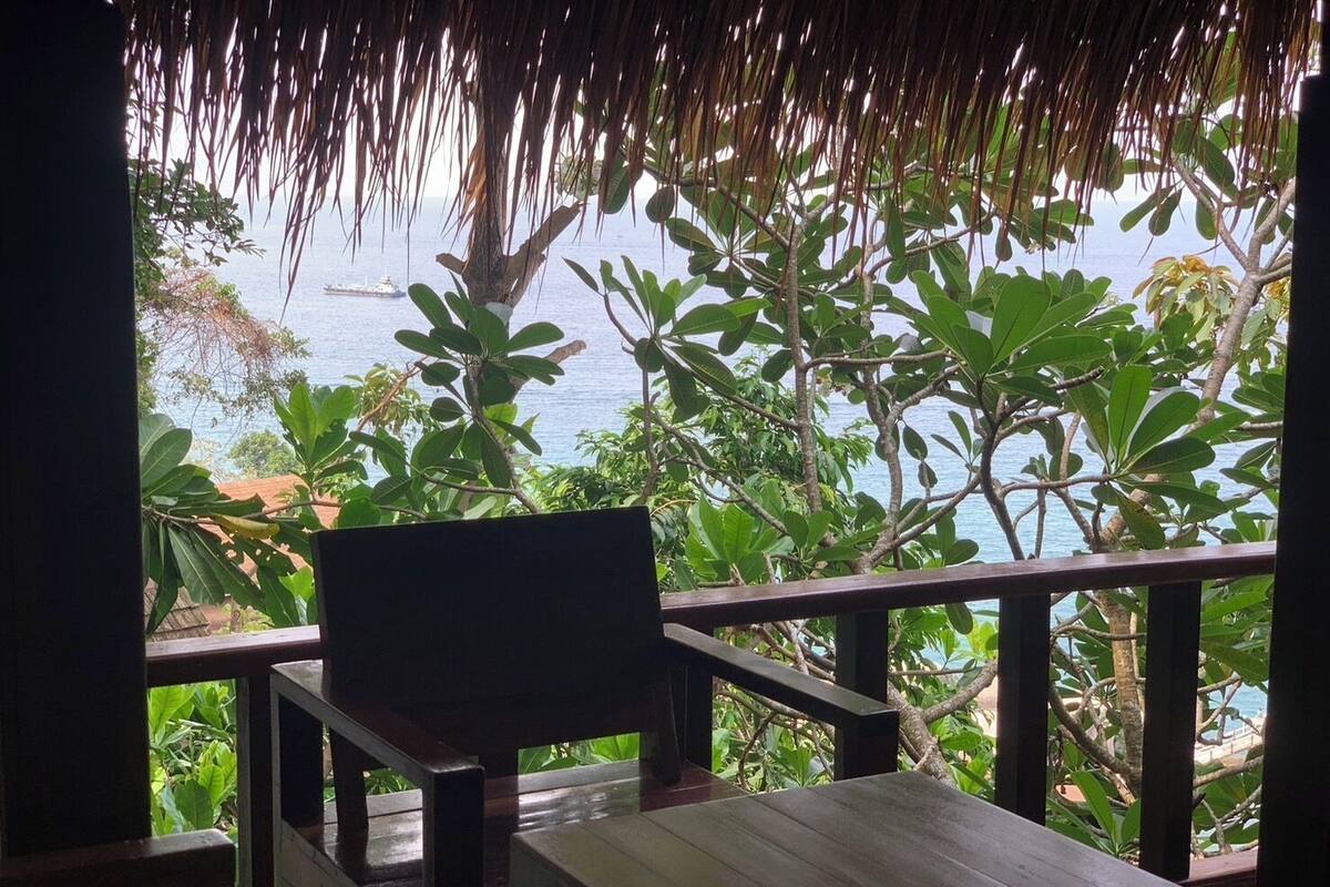 A wooden deck with a thatched roof, overlooking a tropical garden and the sea.