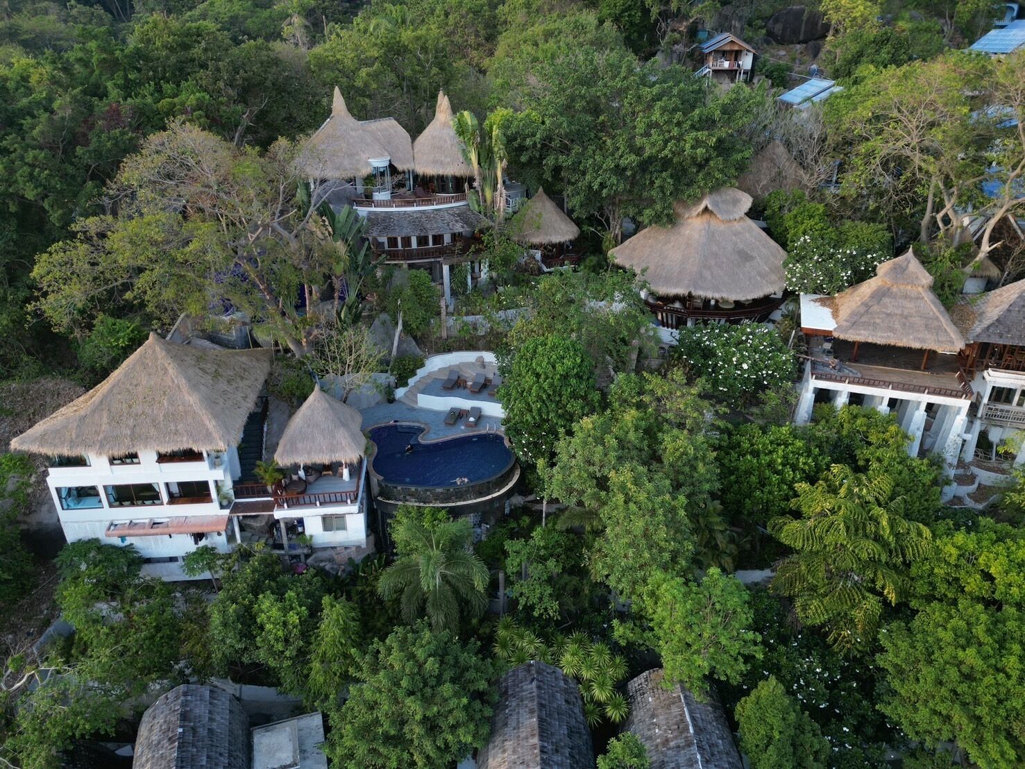 An aerial view of a resort with multiple thatched-roof buildings, a central swimming pool, and lush surrounding vegetation.