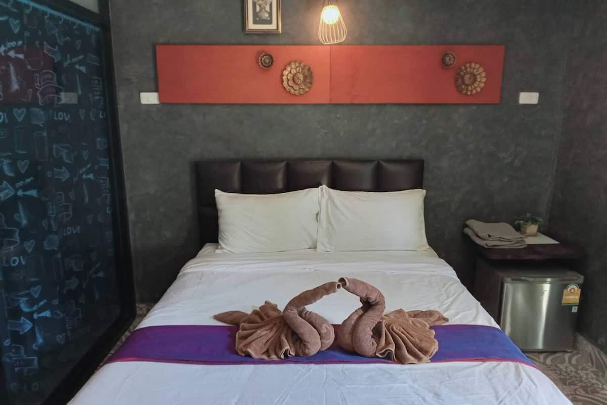 A hotel room with a bed, pillows, a bedside table, a trash bin, and a wall with a shelf and decorative items.