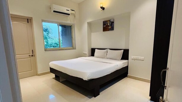 A bedroom with a bed, a ceiling fan, an air conditioning unit, a window, and a door. - Raj Exotic Palms (Vagator)