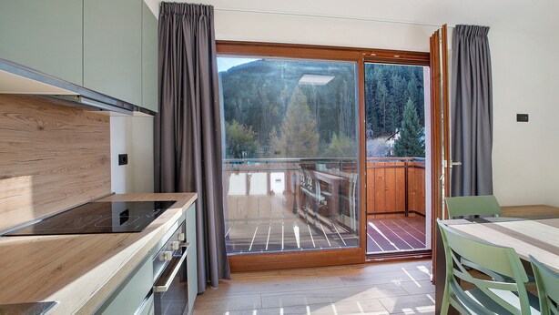 A modern kitchen with a built-in stove, a dining area with a table and chairs, and a sliding glass door leading to a balcony with a view of trees and mountains. - Apartment '3' with Mountain View, Shared Garden and Wi-Fi (Valdidentro)