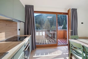 Una cucina moderna con piano cottura integrato, zona pranzo con tavolo e sedie, e una porta scorrevole in vetro che dà su un balcone con vista su alberi e montagne.