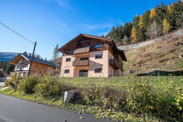 A multi-story building with balconies, a road in the foreground, and a hillside with trees.