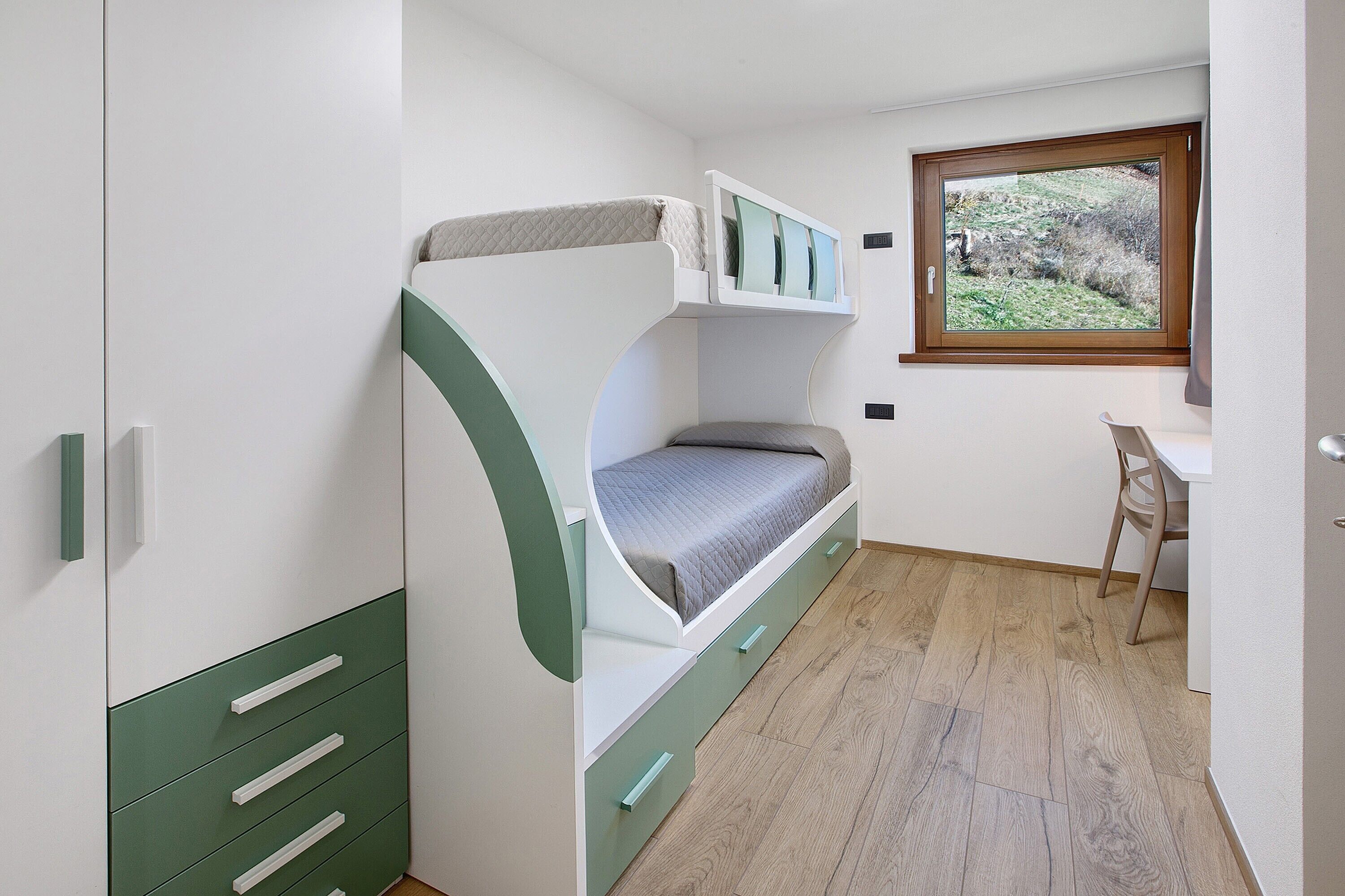 A bunk bed with a desk and chair, a window with a view of greenery, and a wooden floor.