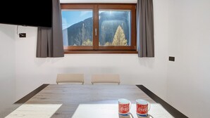 Une pièce dotée d’une grande fenêtre, d’un téléviseur à écran plat fixé au mur, d’une table en bois sur laquelle se trouvent deux canettes de Coca-Cola, et de trois chaises.