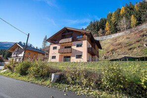 A multi-story building with balconies, a road in the foreground, and a hillside with trees.