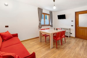 A room with a red sofa, a wooden dining table with red chairs, a TV on the wall, and a door.