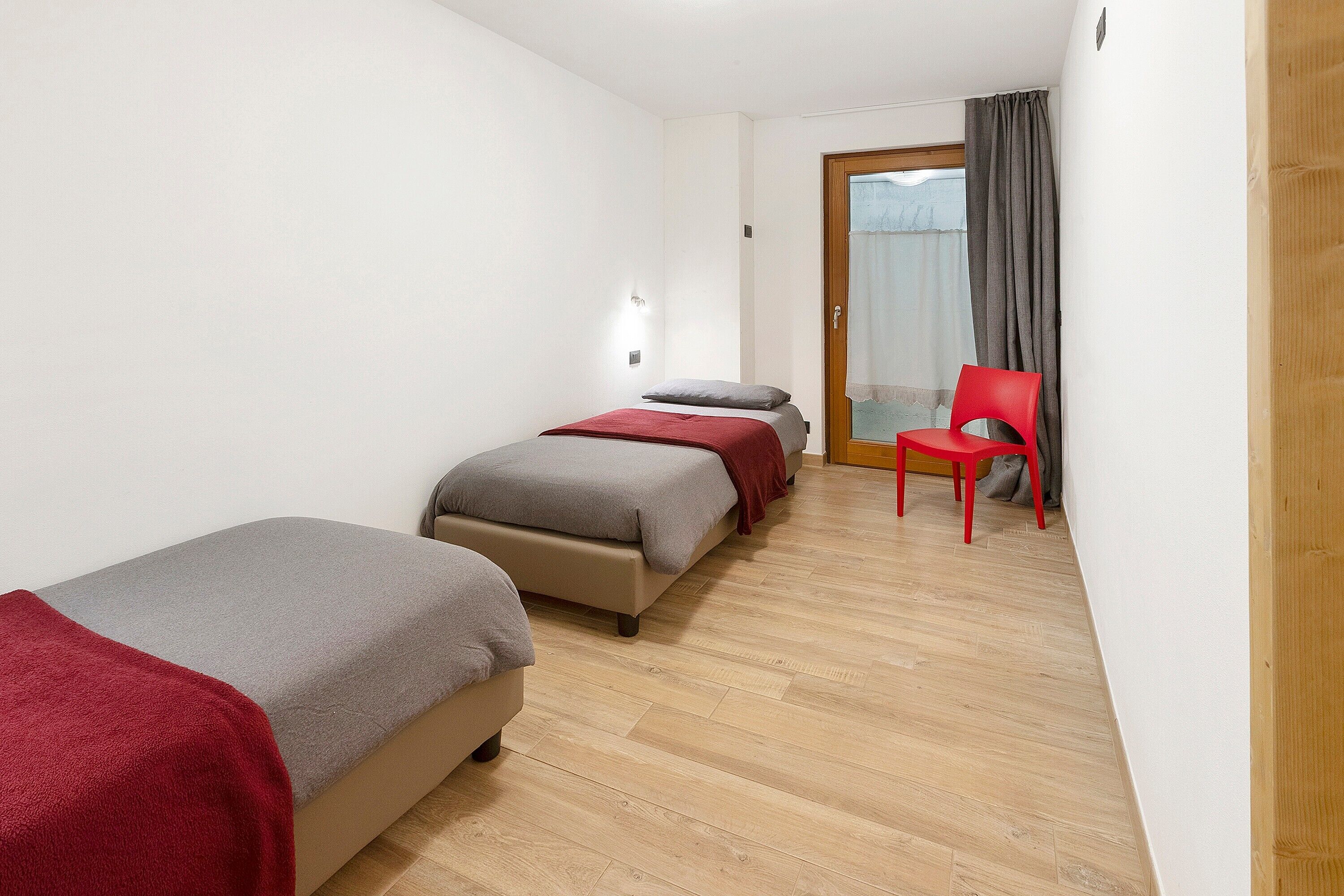 A room with two beds, a red chair, and a wooden floor.