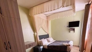 A bedroom with a bed, a nightstand, a chair, a TV, and a wardrobe. - The Victorian Hotel Marsabit (Marsabit)