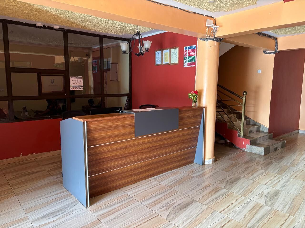 A reception desk with a wooden finish and a grey panel, a red wall with informational posters, and a staircase with a red handrail.