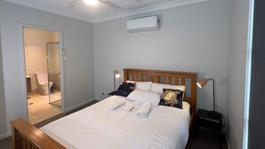 Ein Schlafzimmer mit Bett, Nachttischlampen, Deckenventilator, Klimaanlage und angrenzendem Badezimmer, das durch eine offene Tür sichtbar ist.