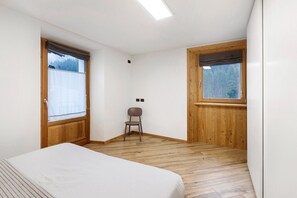 A bedroom with a bed, a chair, wooden flooring, and two windows with curtains.