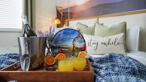 Une chambre d’hôtel avec un lit, une table de chevet avec une lampe, un tableau encadré au mur et un plateau avec une bouteille, des verres et des fruits.