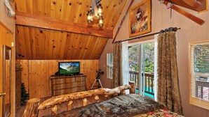 A rustic cabin bedroom with wooden walls, a television on a wooden dresser, a large window with curtains, and a log bench.