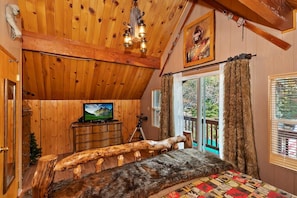 A cabin interior with a wooden ceiling, a television on a dresser, a log bench, and a large window with curtains.
