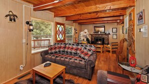 A cozy cabin interior with a wooden ceiling, a fireplace, a sofa with patterned cushions, and a dining area.