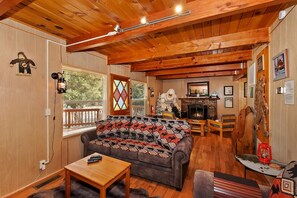 A cozy living room with a wooden ceiling, a sofa with patterned cushions, a fireplace, and a dining area with a table and chairs.