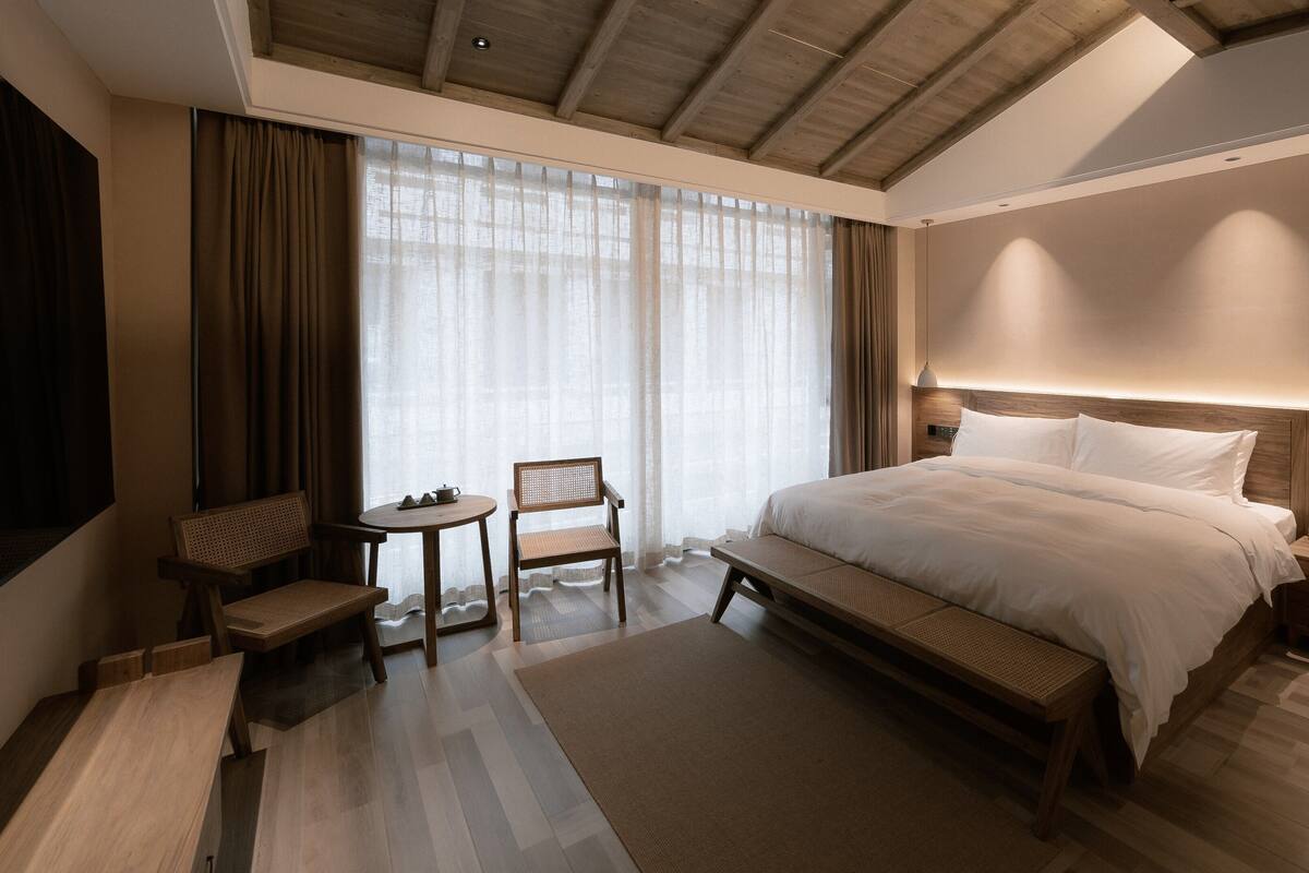 Ein modernes Hotelzimmer mit einem großen Bett, Holzmöbeln, einem Schreibtisch und einem großen Fenster mit Vorhängen.