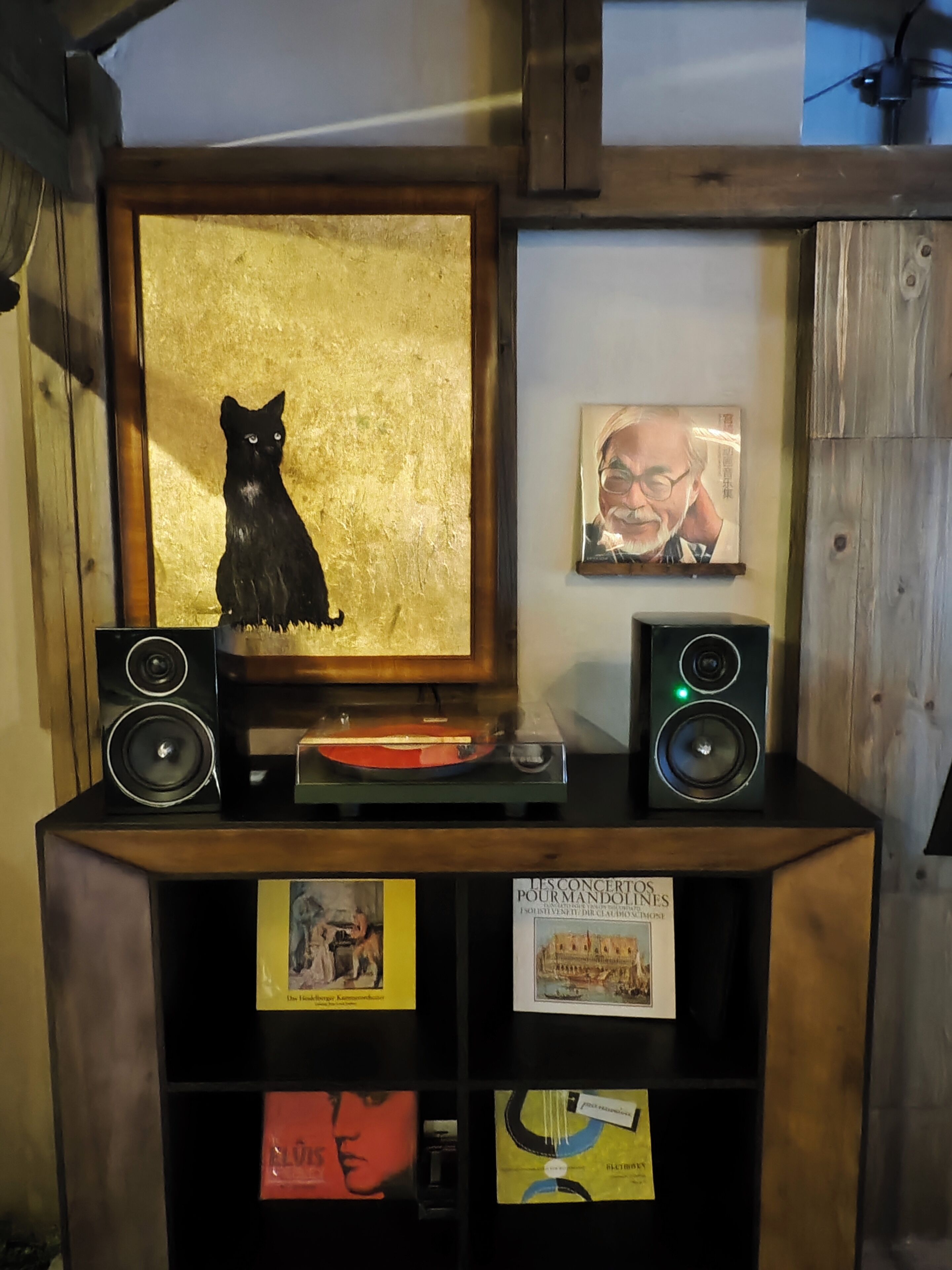 Una estantería de madera con un cuadro enmarcado de un gato negro, dos altavoces y varios objetos pequeños enmarcados.
