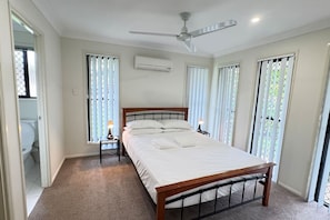 A bedroom with a bed, a ceiling fan, an air conditioning unit, and a window with blinds.