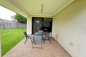 Un patio con mesa y sillas, un espacio cubierto y vista a un jardín trasero.