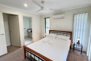 Un dormitorio con una cama, ventilador de techo, aire acondicionado, lámpara de noche y una ventana con persianas.