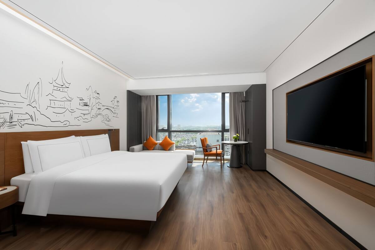 A hotel room with a large bed, a desk, a chair, a TV, and a view of the city.