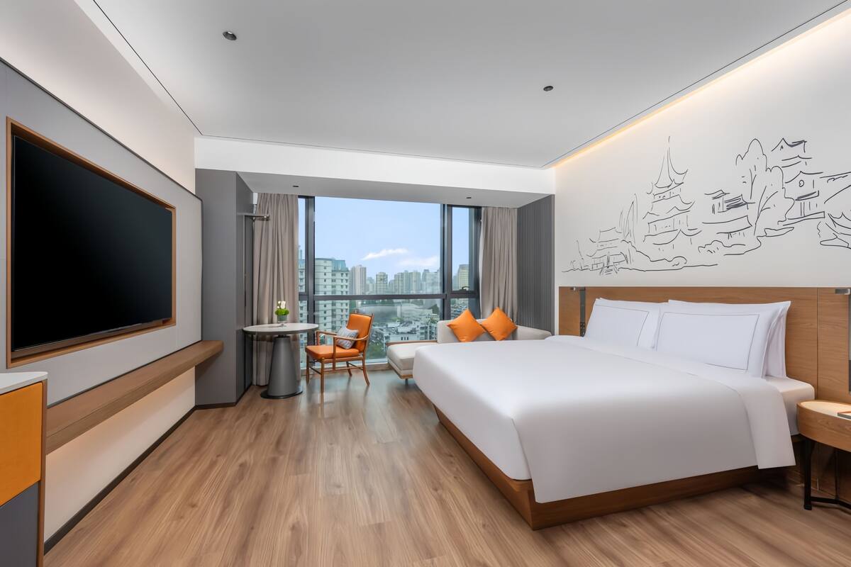 A modern hotel room with a large bed, a desk, a chair, a TV, and a city view.