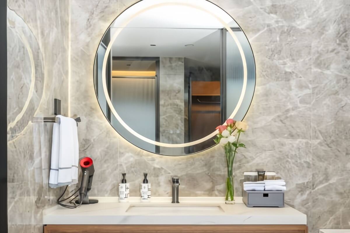 A modern bathroom with a marble wall, a round mirror with a light, a wooden vanity, a towel rack, a hairdryer, a vase with flowers, and a basket.