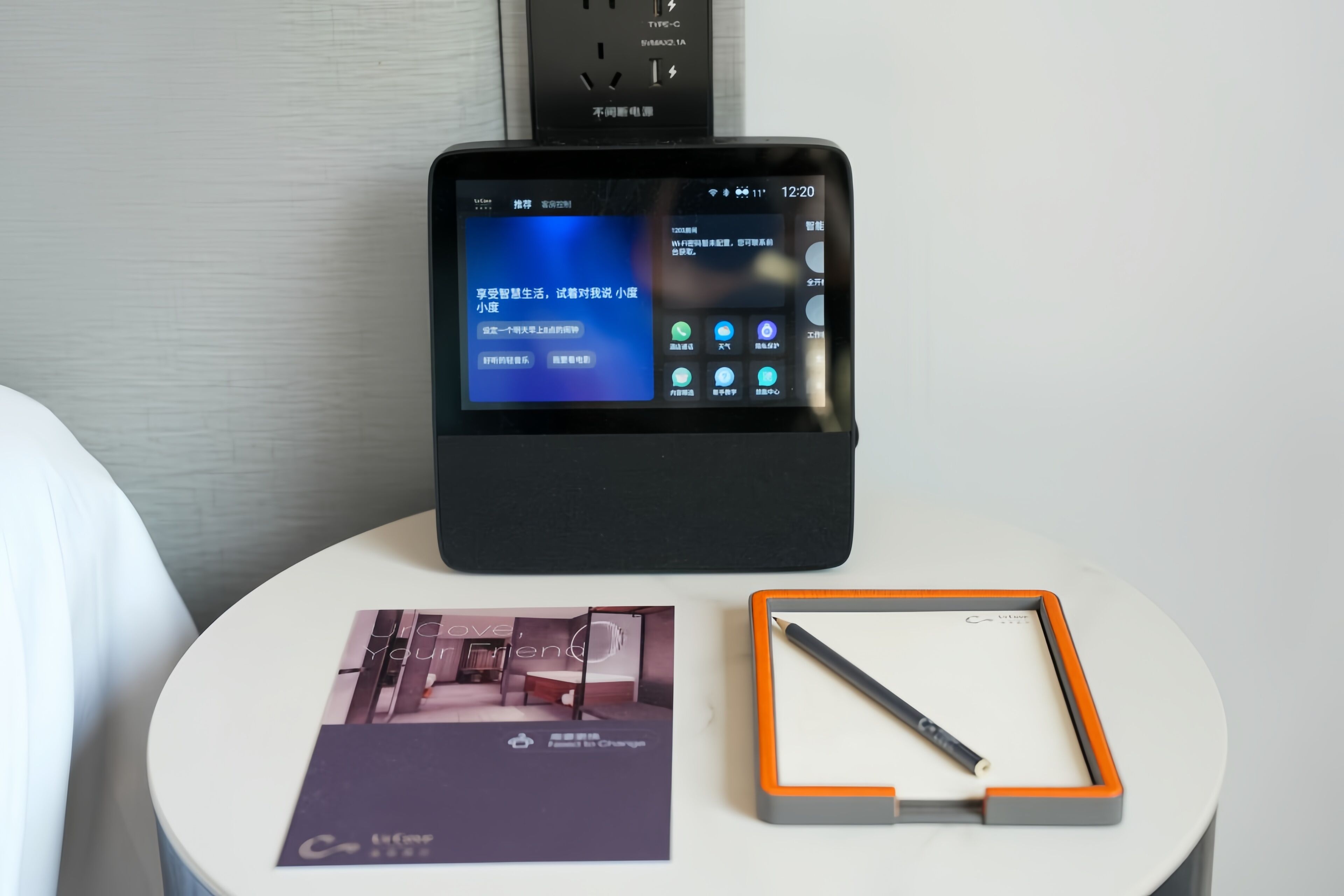 Habitación de hotel con un dispositivo digital en una mesa que muestra un menú, una revista y un bolígrafo en un portalápices.