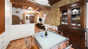 A rustic kitchen with a stone wall, wooden beams, a dining table with two glasses and a bottle, and a cabinet with glassware.