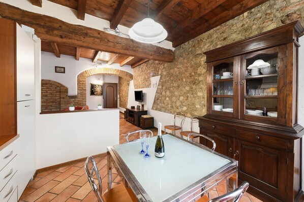 Une cuisine rustique avec un mur en pierre, des poutres en bois, une table à manger avec deux verres et une bouteille, et un buffet contenant de la verrerie.