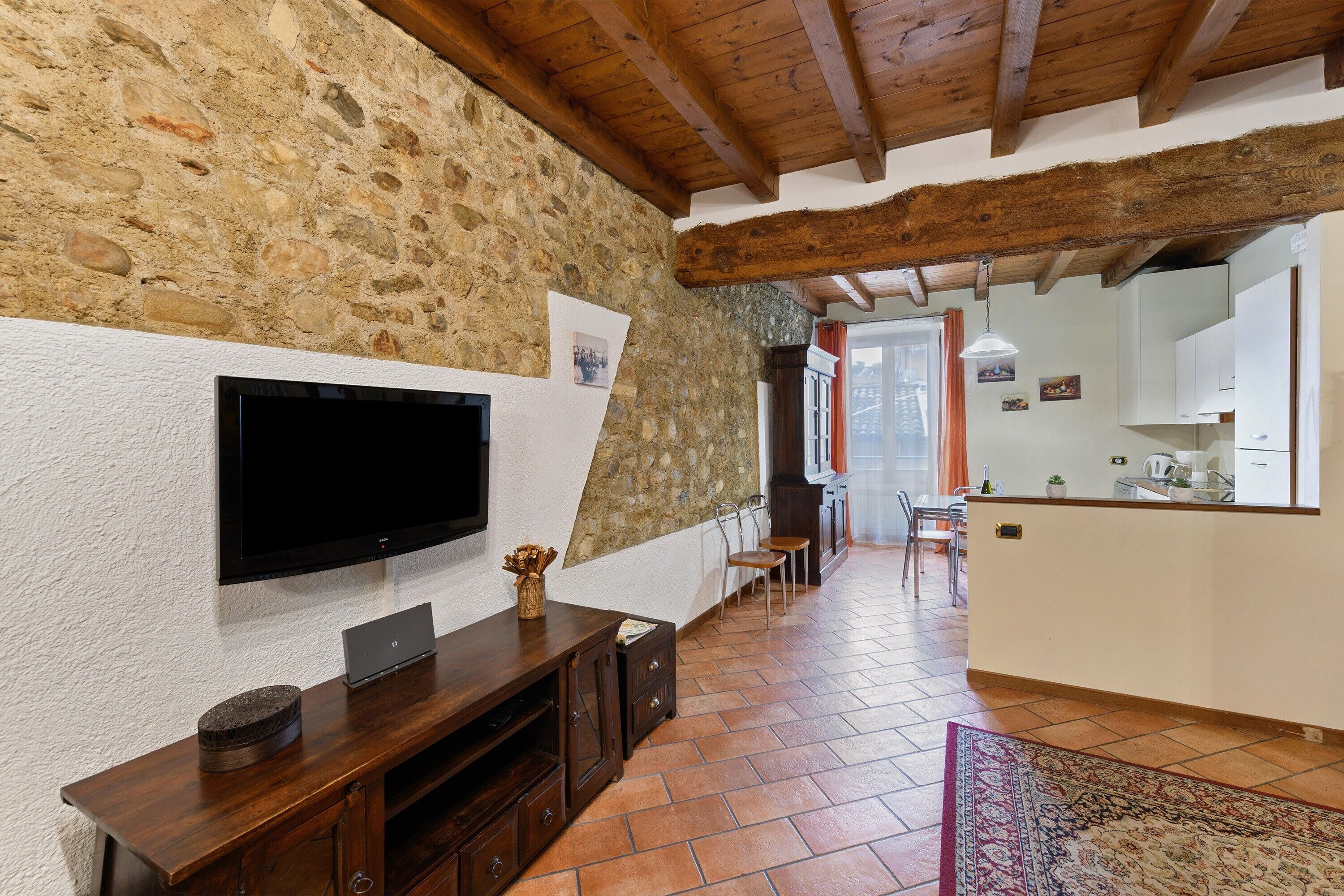 A room with a stone wall, wooden beams, a flat-screen TV, a wooden entertainment unit, a tiled floor, and a kitchen area in the background.
