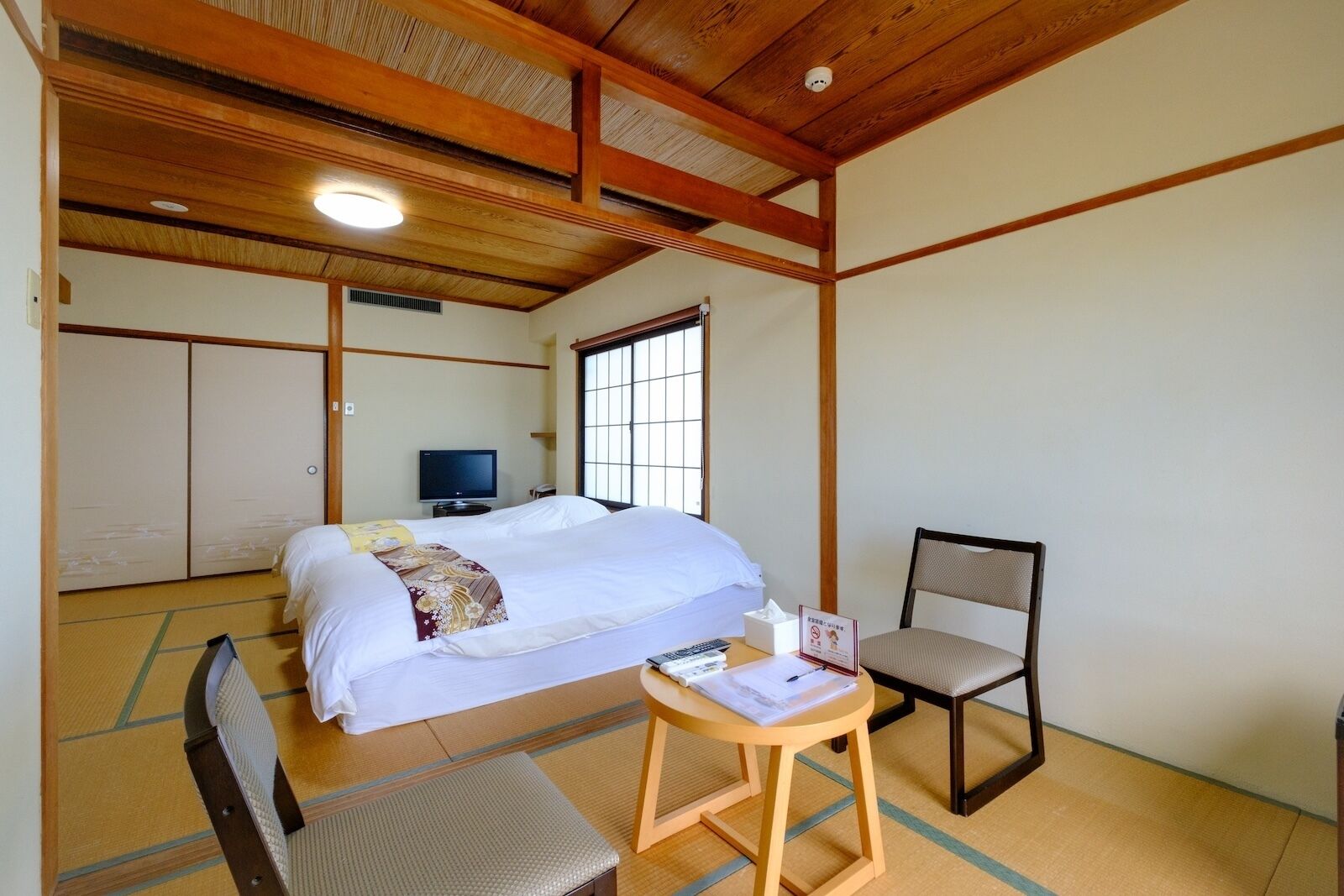 Una habitación tradicional japonesa con suelo de tatami, una cama con colcha blanca, un televisor y una pequeña mesa con una silla.
