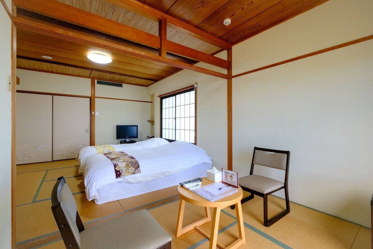 Habitació japonesa tradicional amb sòl de tatami, un llit amb edredó blanc, una televisió i una taula petita amb una cadira.