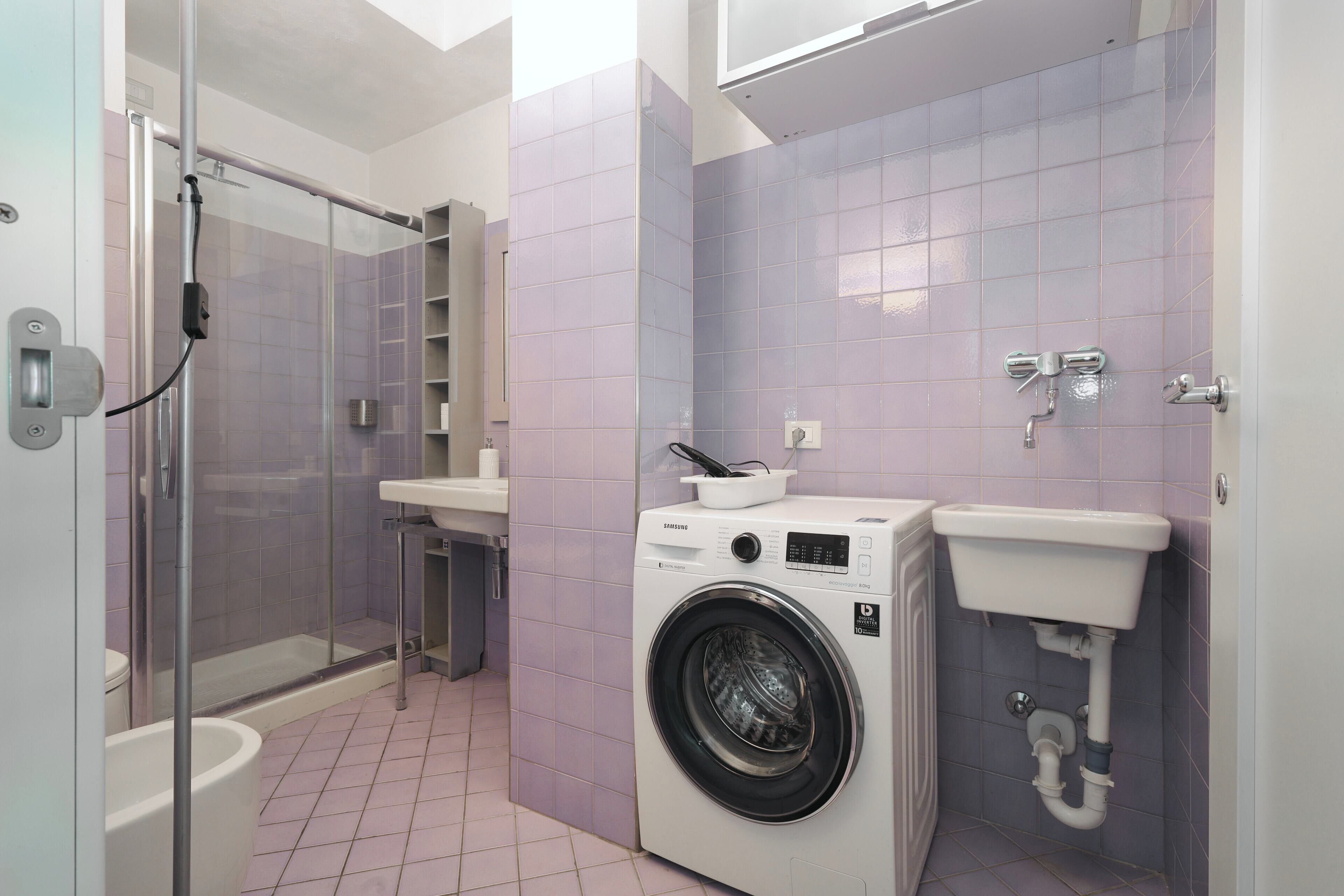 A compact bathroom with a glass shower enclosure, a washing machine, and a sink.