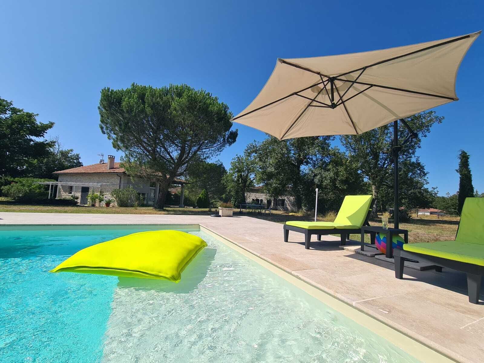 Et poolområde med en klar, blå pool, en gul flydende pude og to grønne liggestole under en beige parasol.