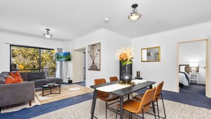 Un salon moderne avec un canapé, une table basse et une table à manger. Une grande fenêtre offre une vue sur les arbres.
