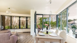 A dining area with a wooden table set for four, large windows offering a view of trees and a balcony with outdoor furniture. - Violino 6 Os b Nowoczesny 5 min do Pla y (Trzebiatow)