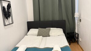 A neatly made bed with a teal and beige bedspread, two pillows, and two rolled towels. A black headboard, a small bedside table with a lamp, and a framed leaf decoration on the wall are also visible.