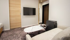 Hotelkamer met een houten deur, een flatscreen televisie aan de wand, een klein tafeltje met een afstandsbediening, een zwarte printer en een beige bank.