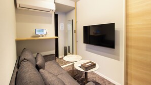Een moderne hotelkamer met een grijze bank, een klein rond tafeltje, een flatscreen televisie aan de muur en een airconditioning.