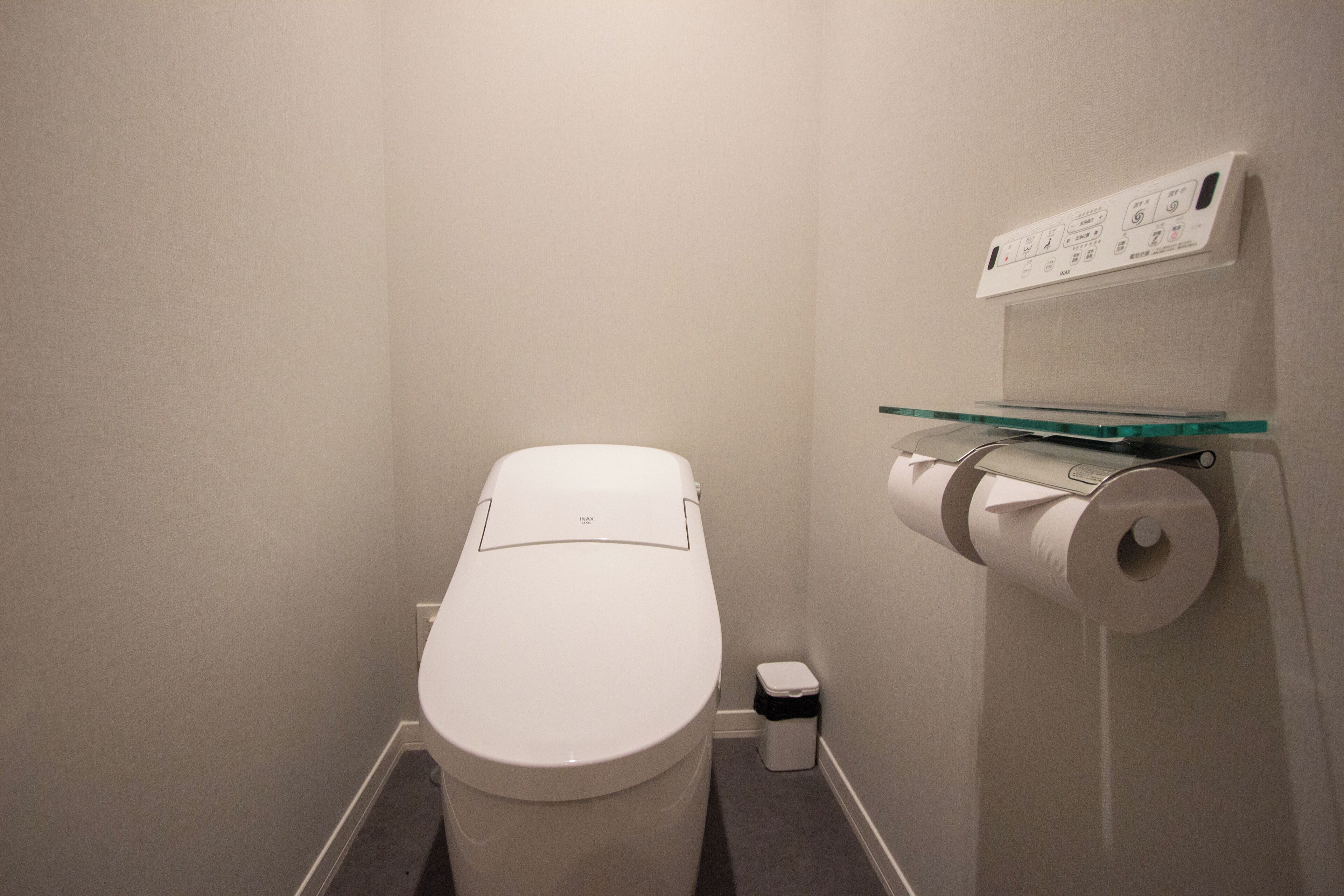 A small, modern bathroom with a white toilet, a glass shelf holding a toilet paper roll, and a trash bin.