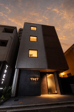 Un bâtiment moderne doté d’un toit plat, de trois fenêtres illuminées et d’une enseigne sur laquelle on peut lire « TEET TENJIN-MINAMI ».