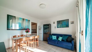 Una sala de estar moderna con un sofá azul, una mesa de comedor con sillas, una zona de cocina y un amplio ventanal con cortinas.