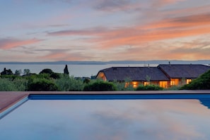 Une piscine avec vue sur une maison et un plan d’eau, au coucher du soleil.